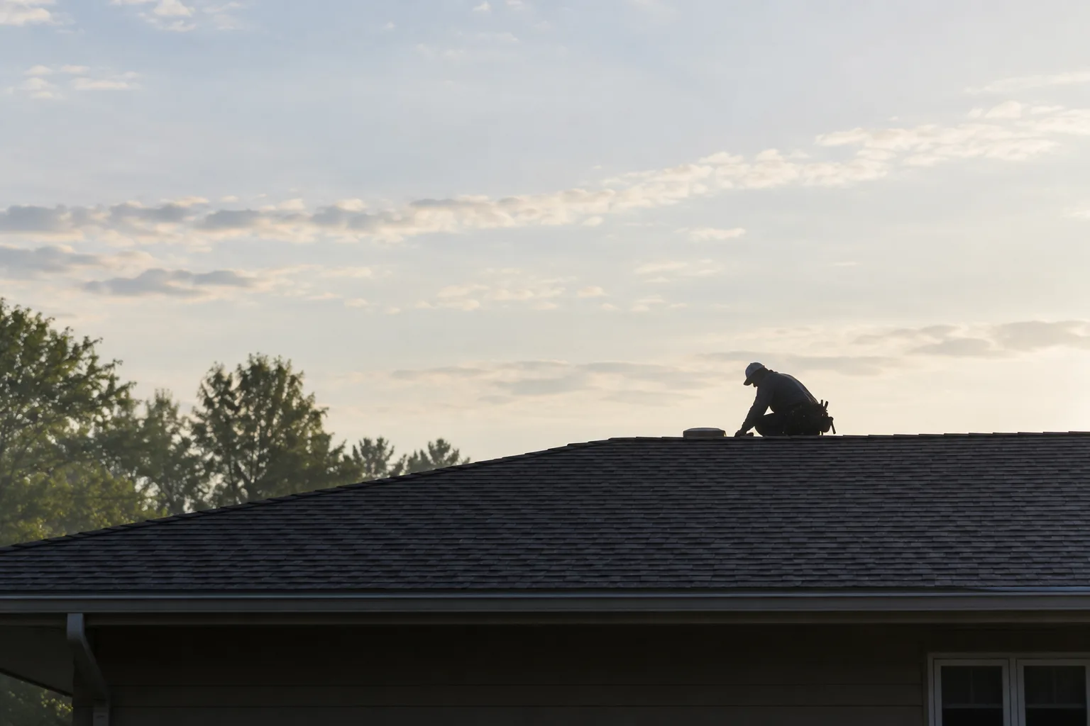 Backlit silhouette of a crouching roofer above a shingle roof against a pale morning sky